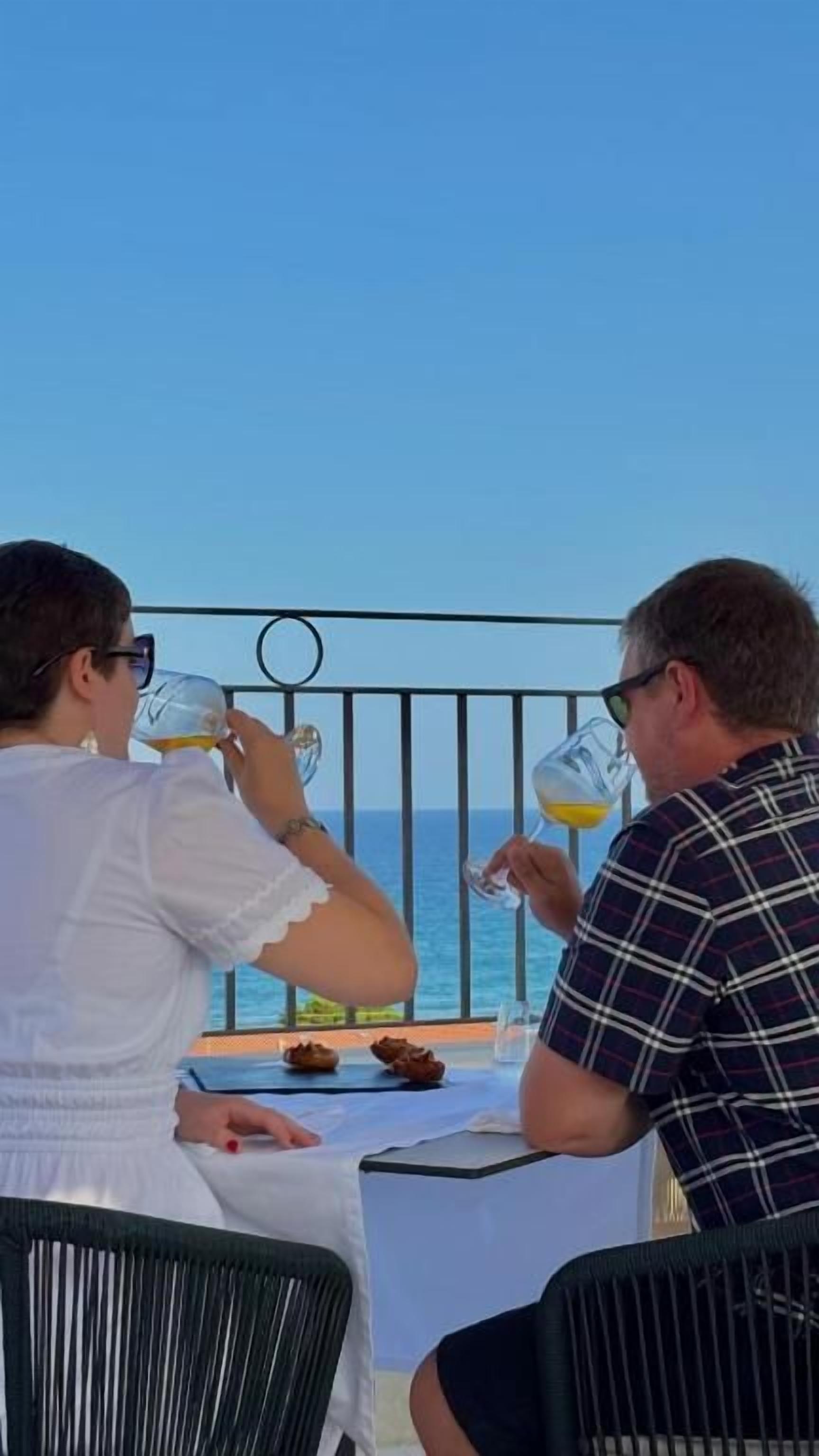 Two people seated at a table with a view of the sea, a metal railing, and a tablecloth.