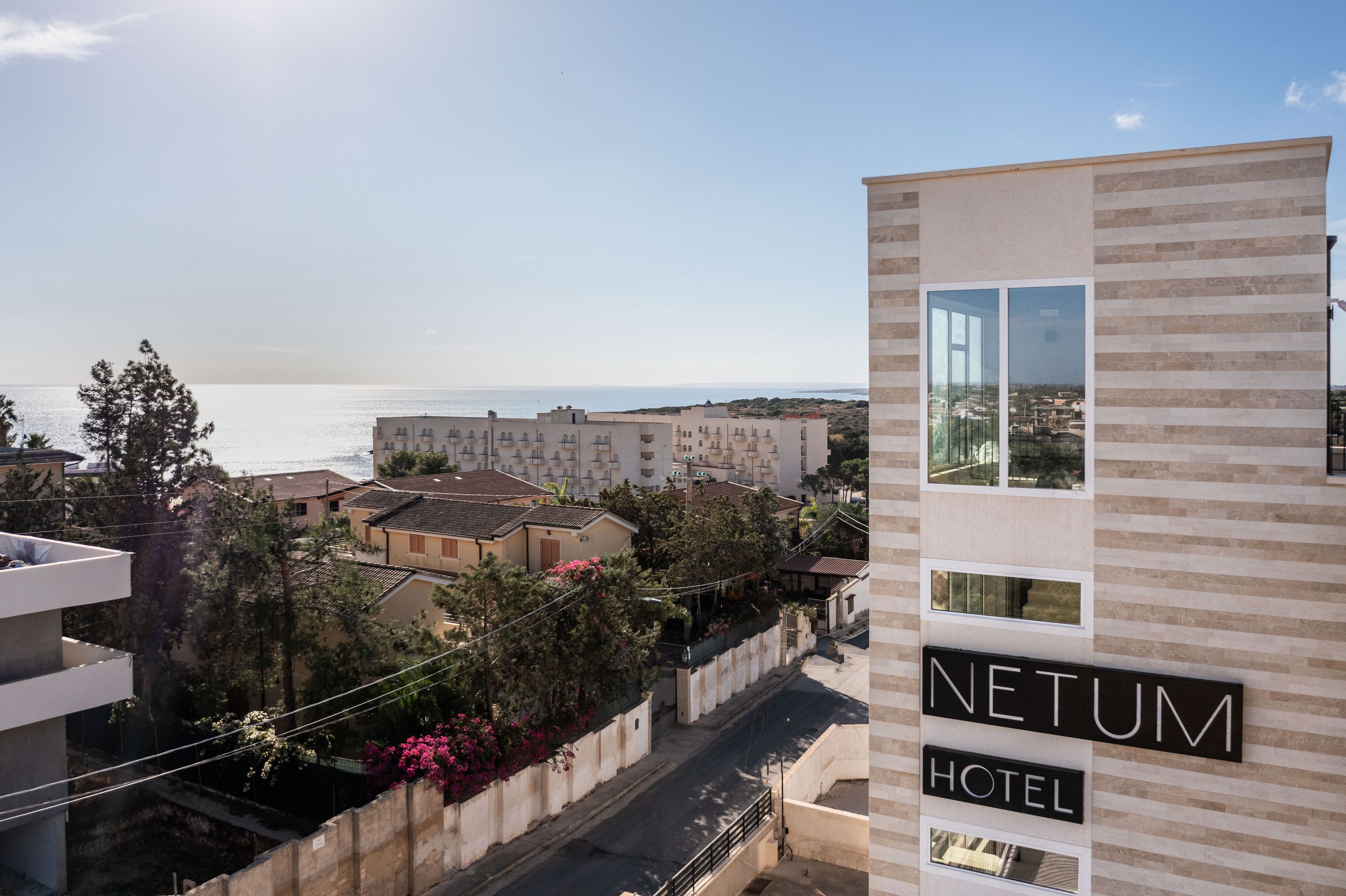 Un bâtiment d’hôtel moderne, avec une enseigne indiquant « NETUM HOTEL », et une vue sur la mer en arrière-plan.