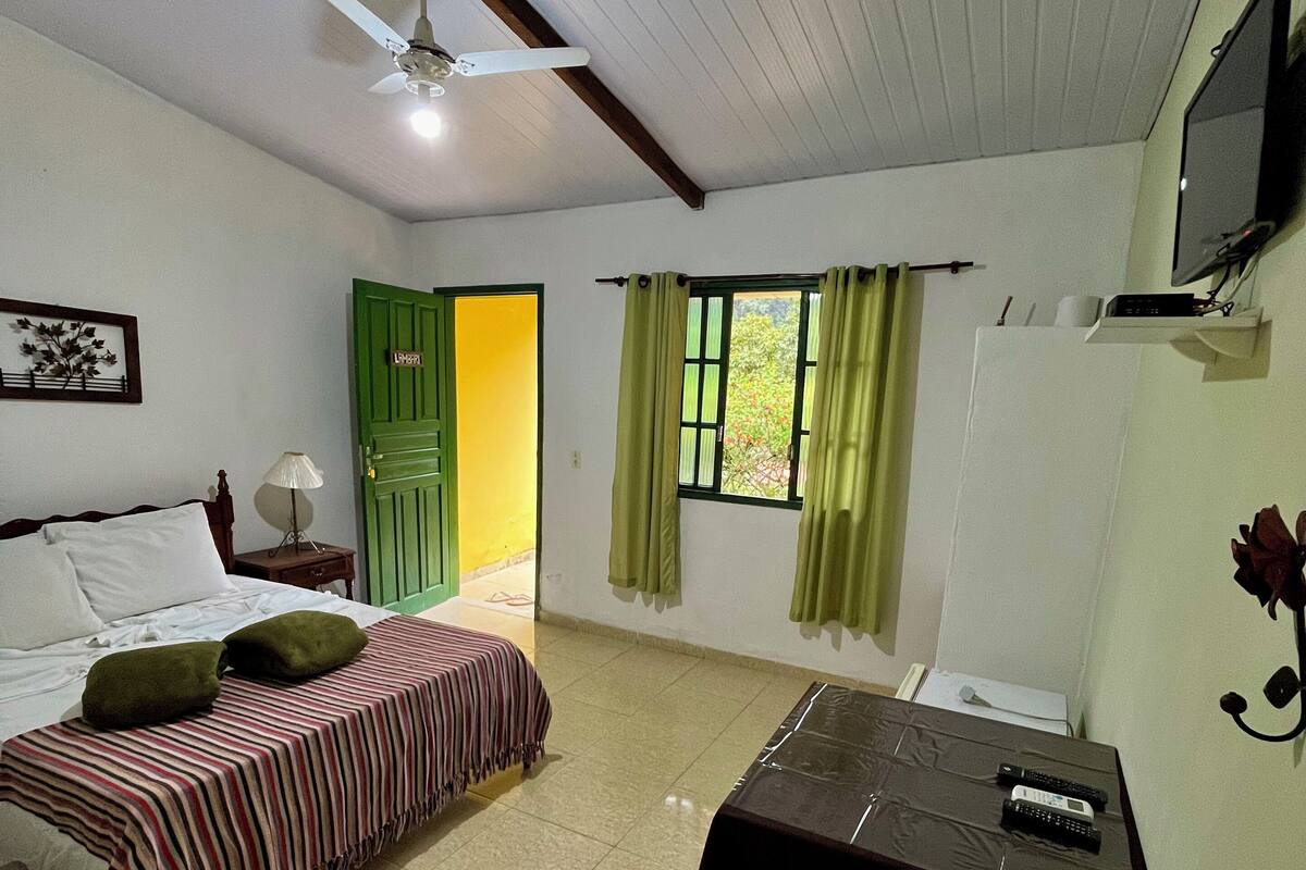 Ein Zimmer mit einem Bett, einem Schreibtisch, einem Sessel, einem Deckenventilator, einem Fenster mit Vorhängen und einem an der Wand befestigten Fernseher.
