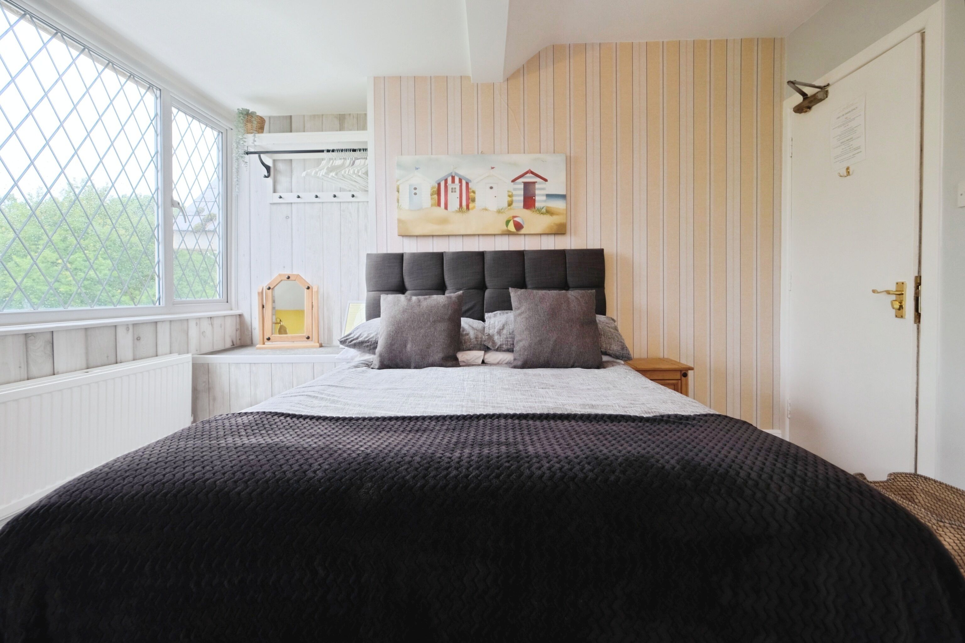 A bedroom with a large bed, a headboard, a painting on the wall, and a window with a view of greenery.