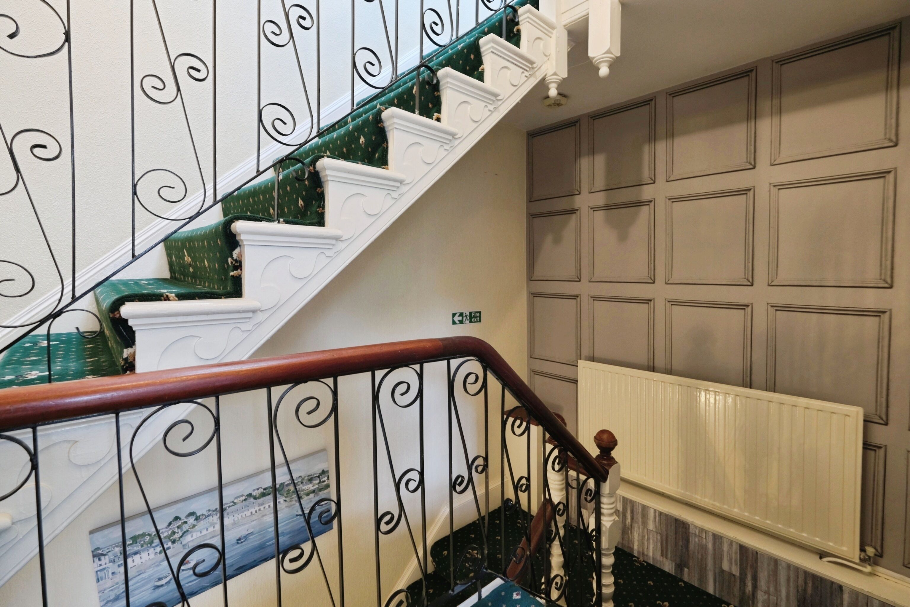 A staircase with ornate black metal railings and white steps, leading to a landing with a white radiator and a wall with square panels.