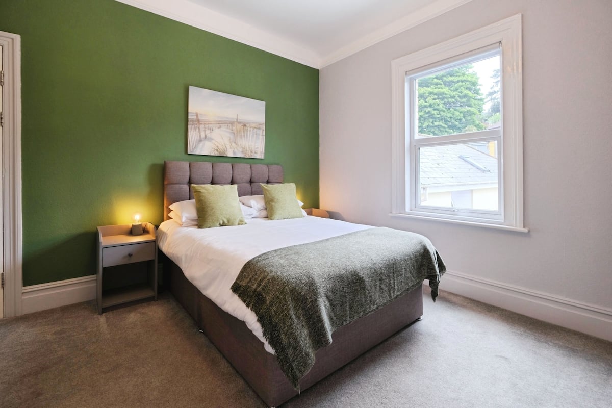 A bedroom with a bed, green walls, a window, and a nightstand with a lamp.
