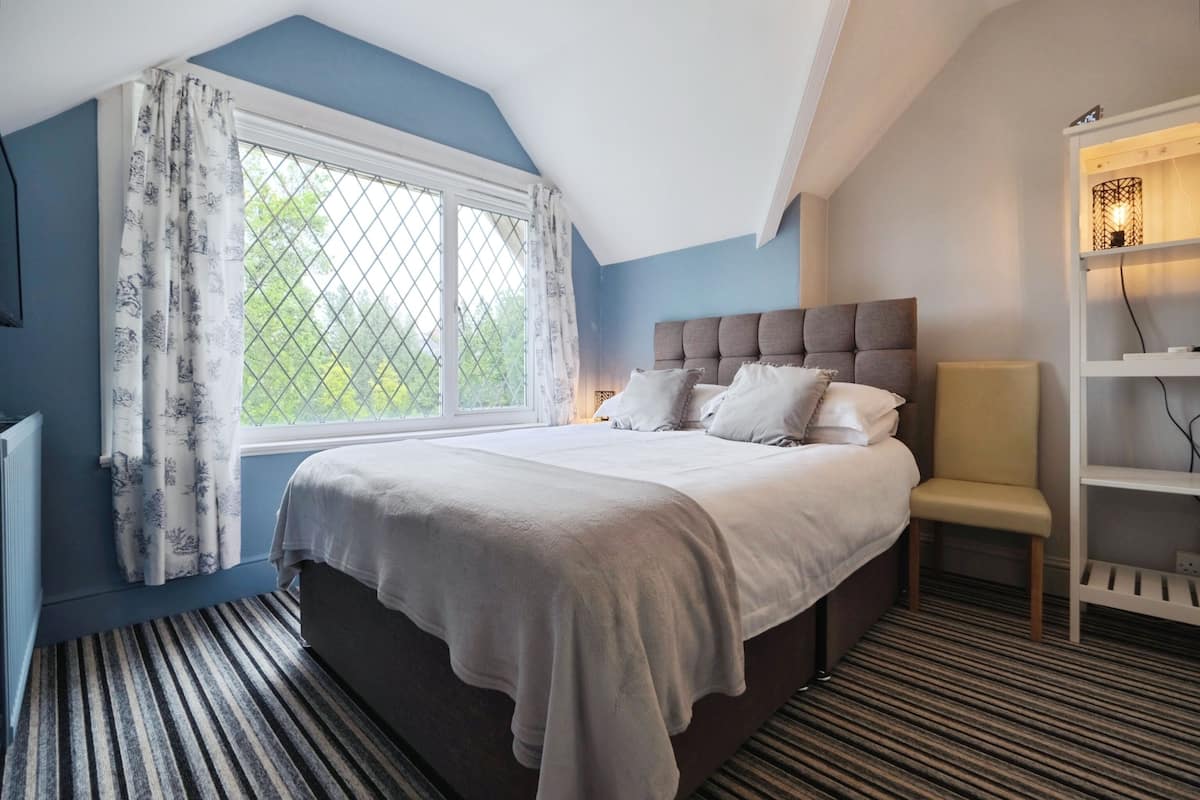 A bedroom with a large bed, a window with curtains, a bedside table, a chair, and a shelf with a lamp.