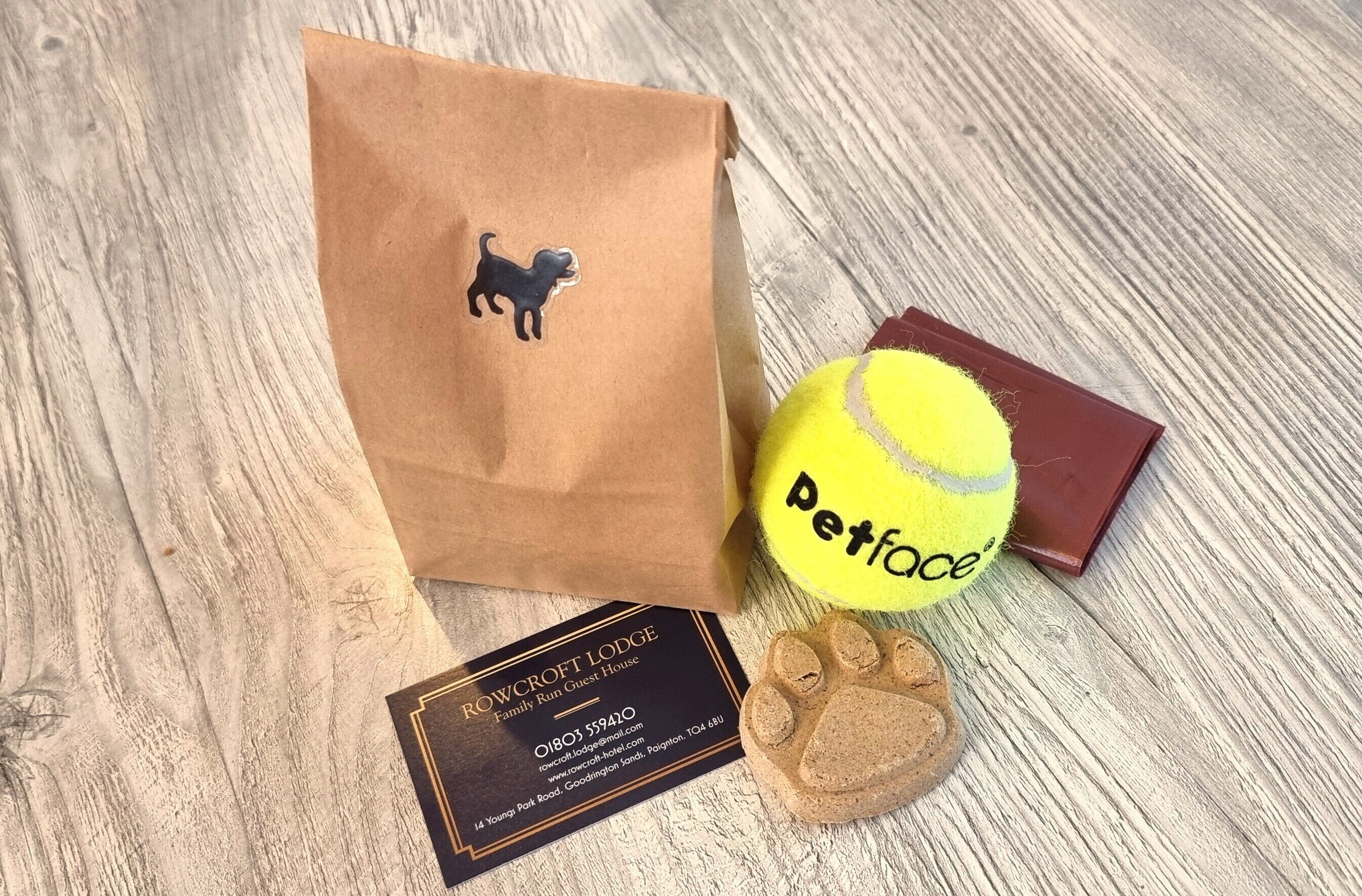 A brown paper bag with a dog logo, a yellow tennis ball with "Petface" written on it, a brown paw print, and a business card for Rowcroft Lodge.
