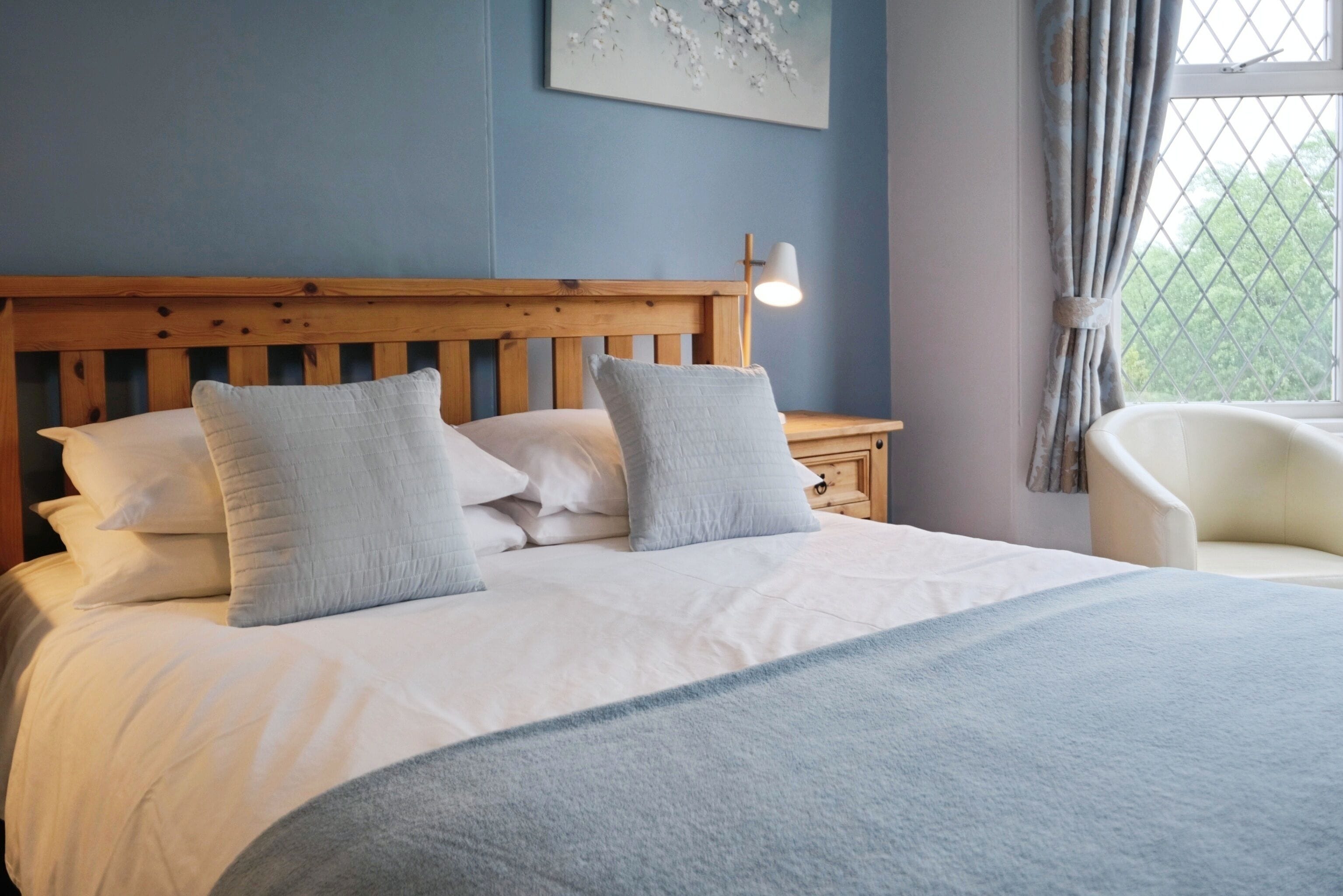 Una cama bien hecha con sábanas blancas y dos almohadas, un cabecero de madera, una mesita de noche con una lámpara y una ventana con cortinas.