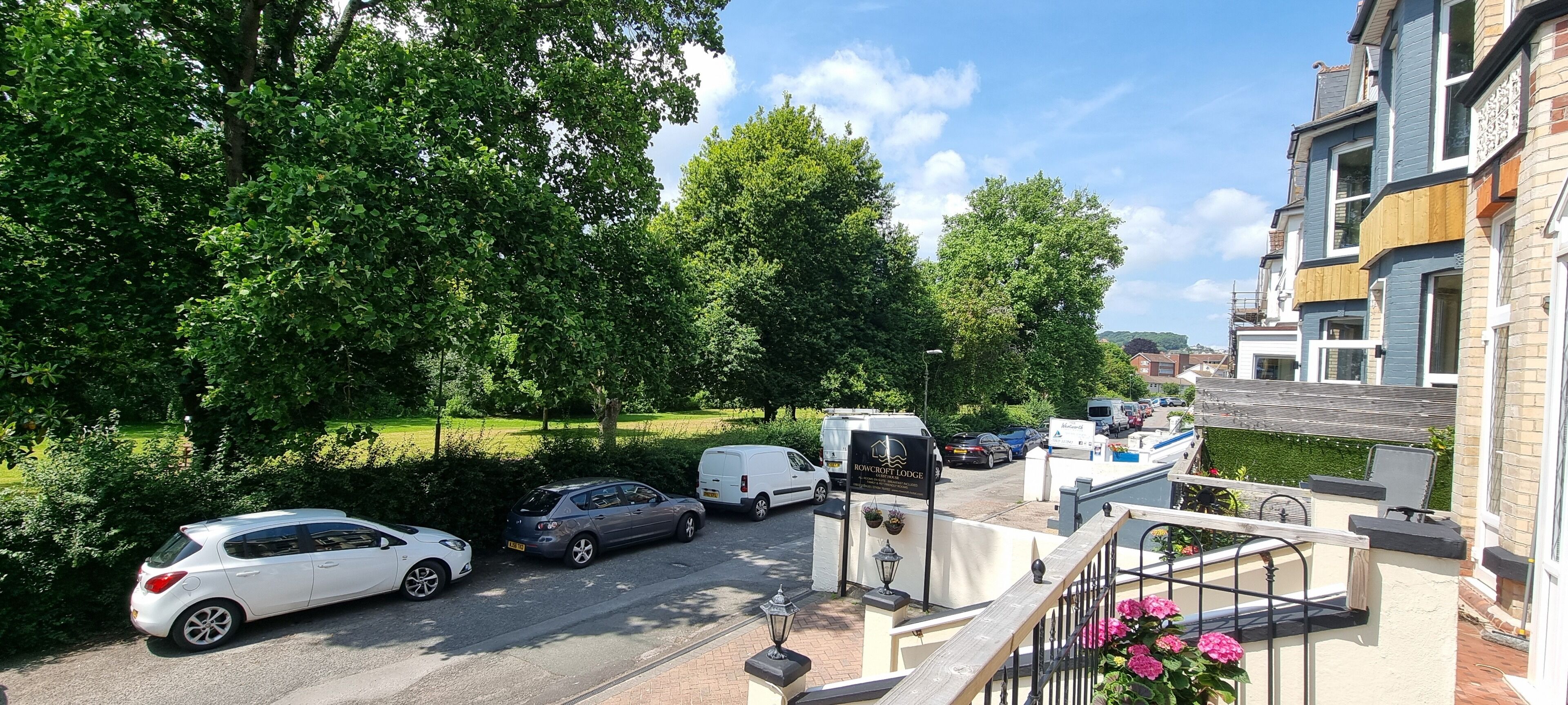 A parking area with several cars and a white van, surrounded by lush greenery and a building with a balcony.