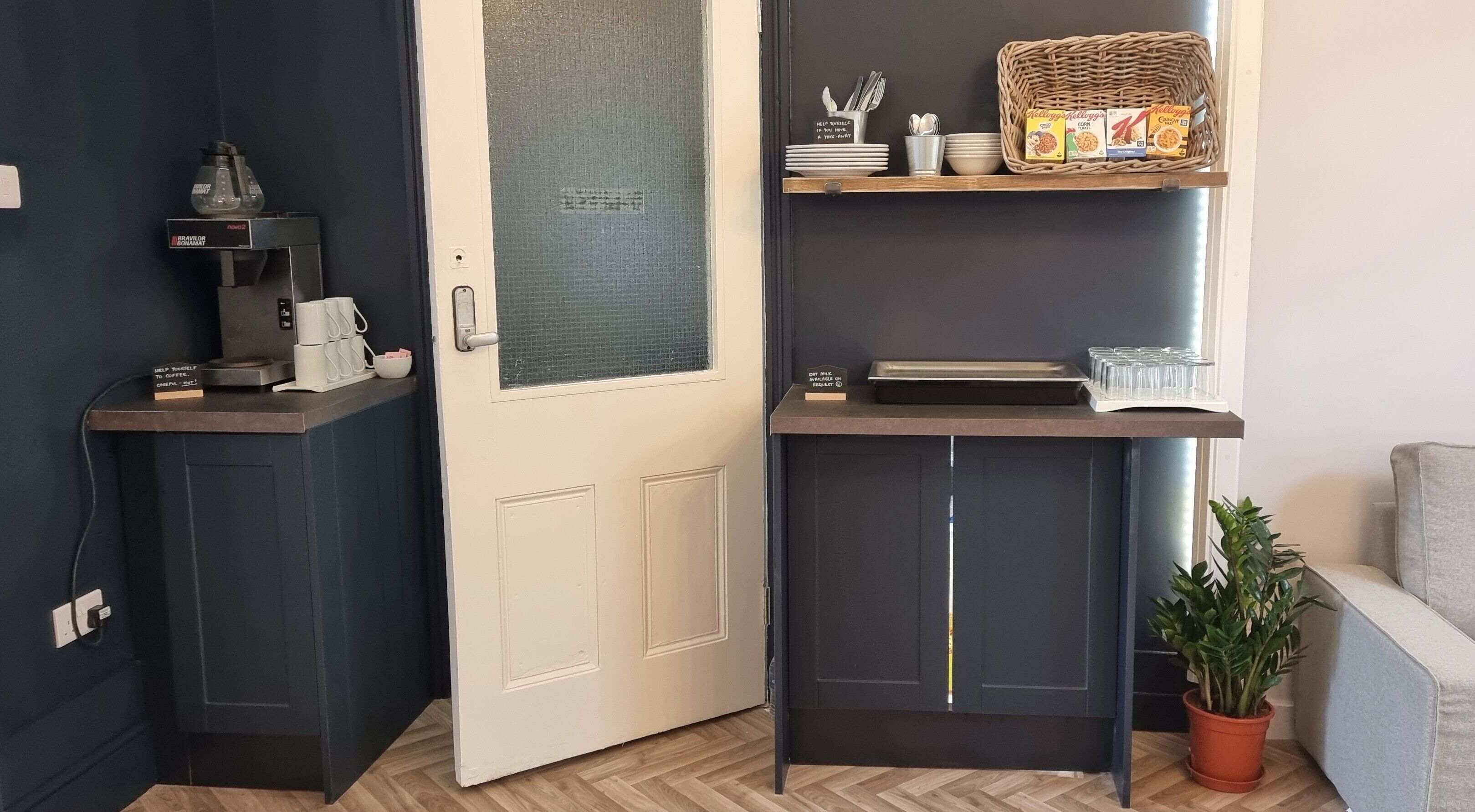 A compact room with a door, a coffee machine, a shelf with breakfast items, and a potted plant.