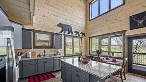 A modern kitchen with granite countertops, grey cabinets, and a large island. There are multiple windows providing natural light and views of the outdoors.