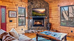 Un interior de cabaña acogedor, con chimenea de piedra, paredes de madera, televisor y una zona de estar confortable.