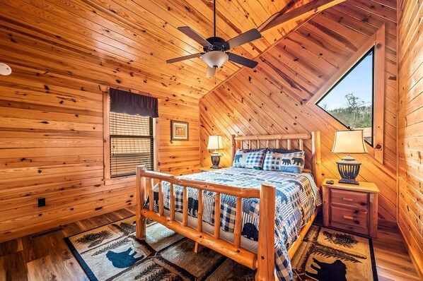 Une chambre confortable dans un chalet en rondins, avec un plafond en bois, un lit recouvert d’une couette bleue et blanche, deux lampes de chevet et une fenêtre donnant sur le paysage extérieur.