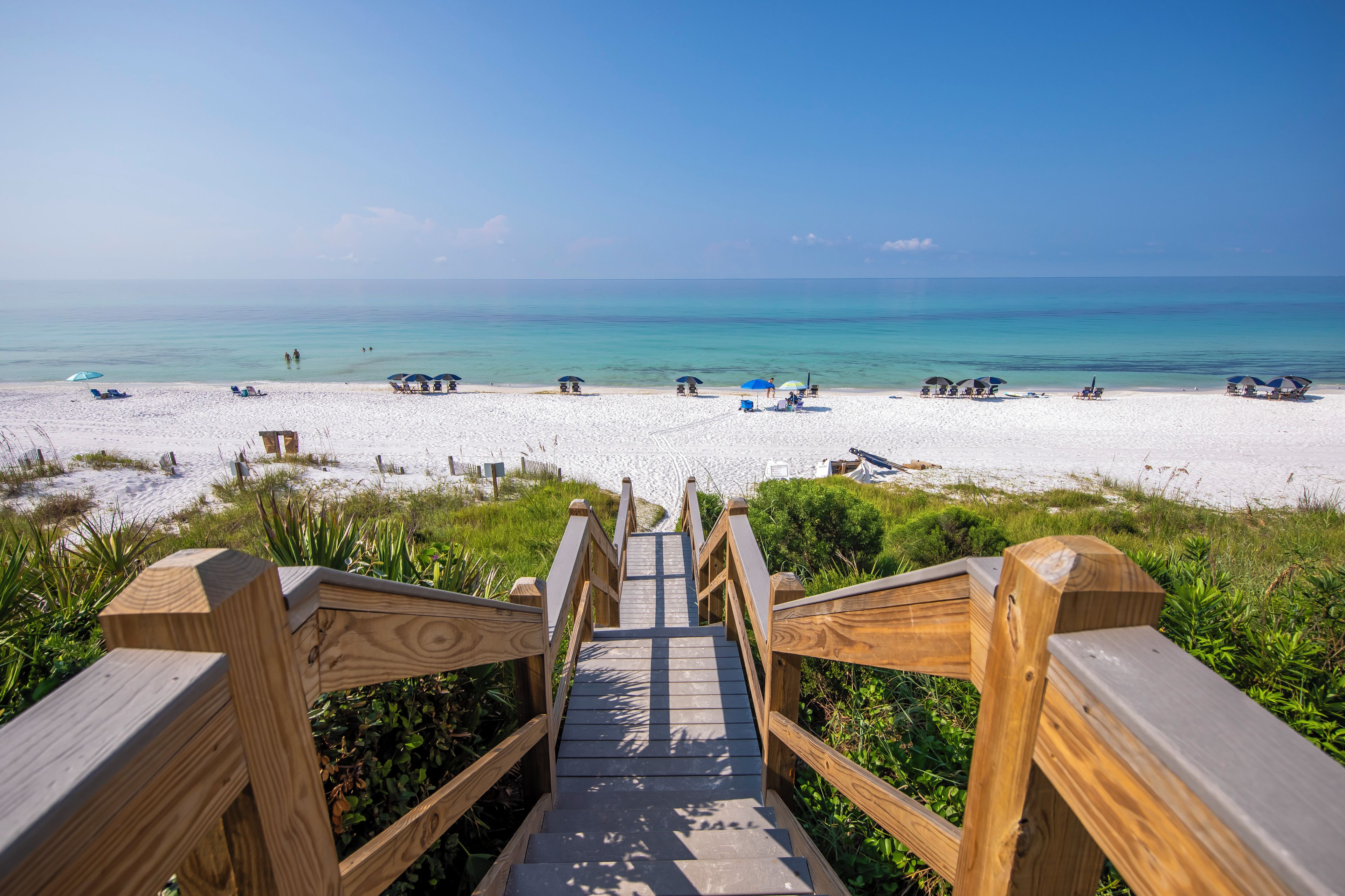 Eine Holztreppe führt zu einem Strand mit weißem Sand und klarem, blauem Wasser.