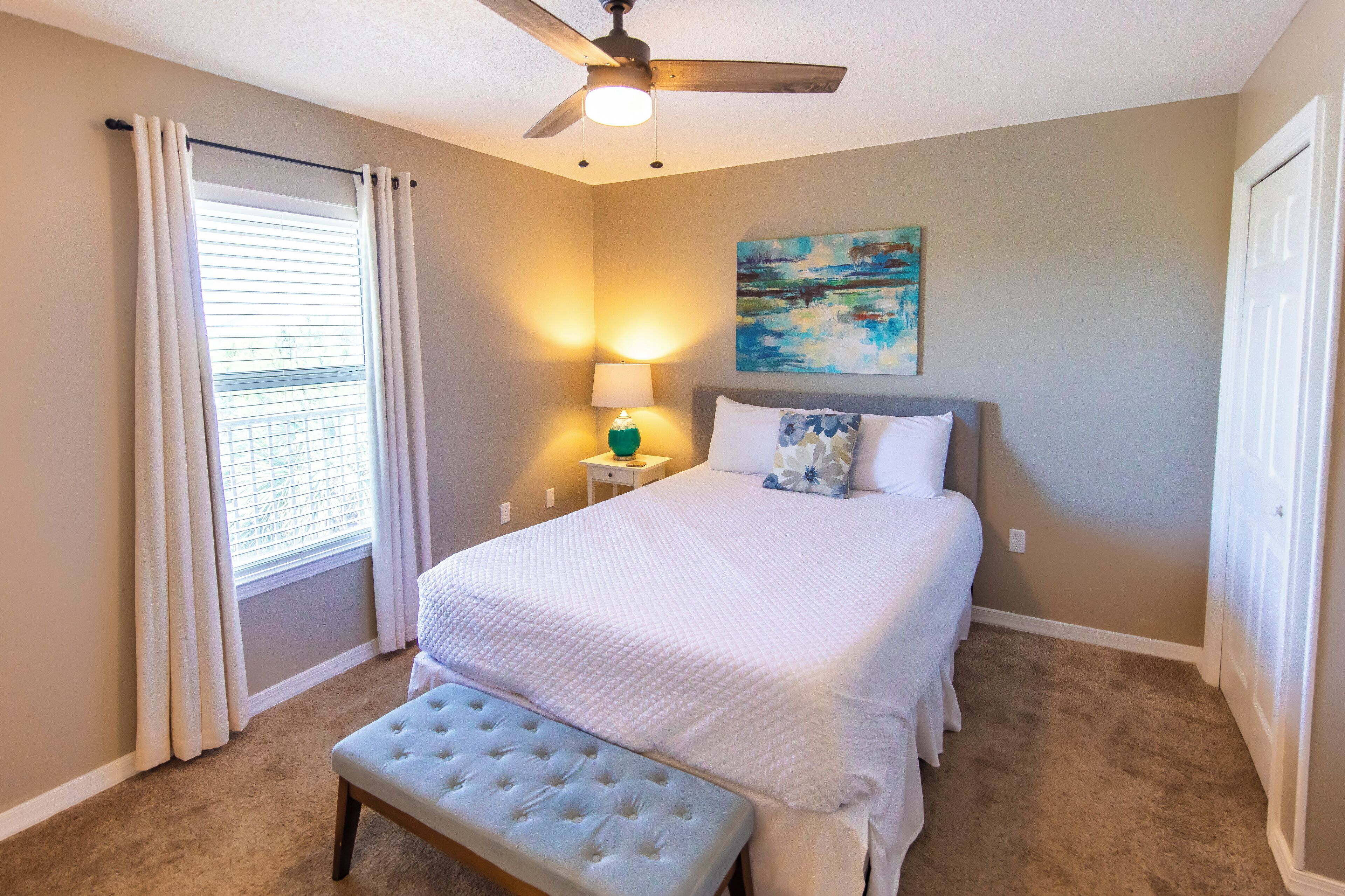 A bedroom with a bed, a bench, a ceiling fan, a window with curtains, a nightstand with a lamp, and a painting on the wall.