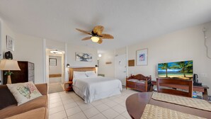 Ett hotellrum med en säng, ett skrivbord, en stol, en soffa, en takfläkt och en tavla som föreställer en strandvy.