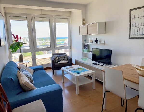 Una sala de estar moderna con un sofá azul, una mesa de centro blanca, un televisor y un balcón con vistas a la ciudad.
