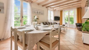 Un coin repas avec une table dressée pour quatre personnes, un canapé et un téléviseur, dans une pièce lumineuse dotée de poutres en bois et de grandes fenêtres.