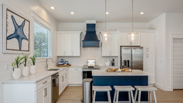 A modern kitchen with white cabinetry, a blue island, and a stainless steel refrigerator. There are two white stools at the island and a hanging light fixture above. - Mariner: New! Private Beach! (Miramar Beach)