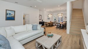A spacious living room with a white sofa, a coffee table, and a dining area with a wooden staircase. - Mariner: New! Private Beach! (Miramar Beach)