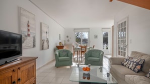 Una sala de estar con televisor, dos sillones verdes, una mesa de centro de vidrio con fruta, un sofá color beige y un comedor al fondo.