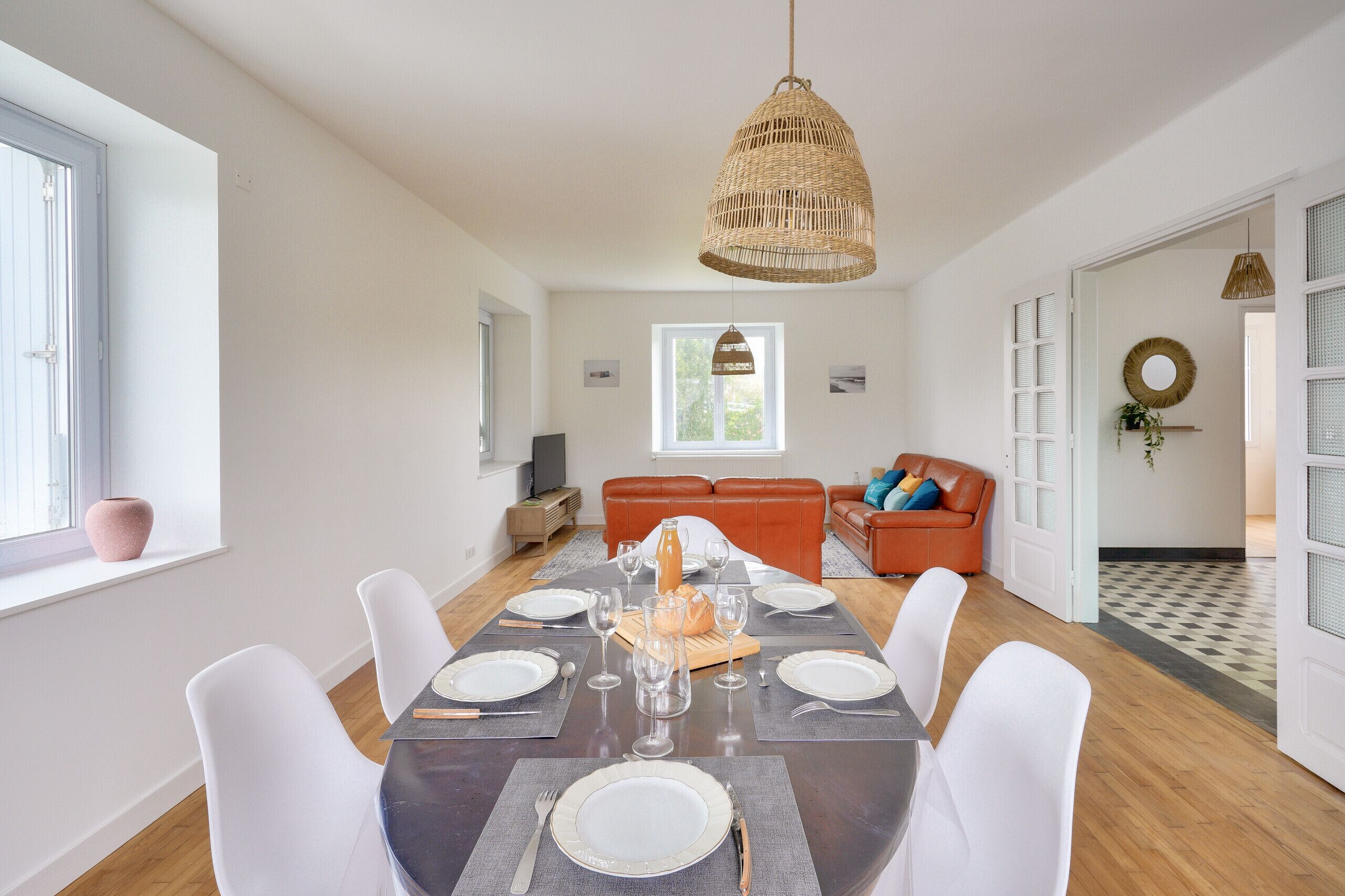 A dining area with a table set for six, a wicker pendant light, and a view of a living room with a sofa and a plant.
