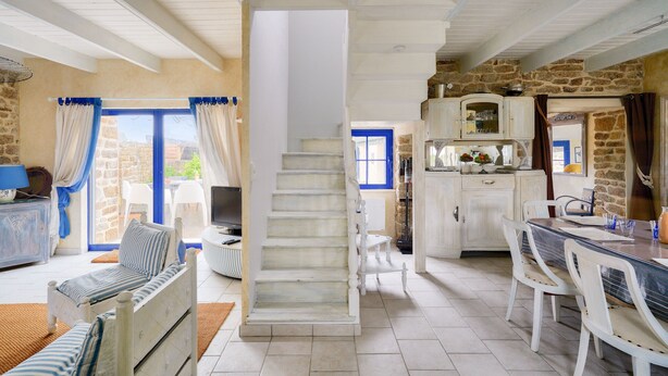 Un vaste espace de vie comprenant un escalier, une table à manger avec des chaises, et une cuisine avec des murs en pierre et des meubles blancs.