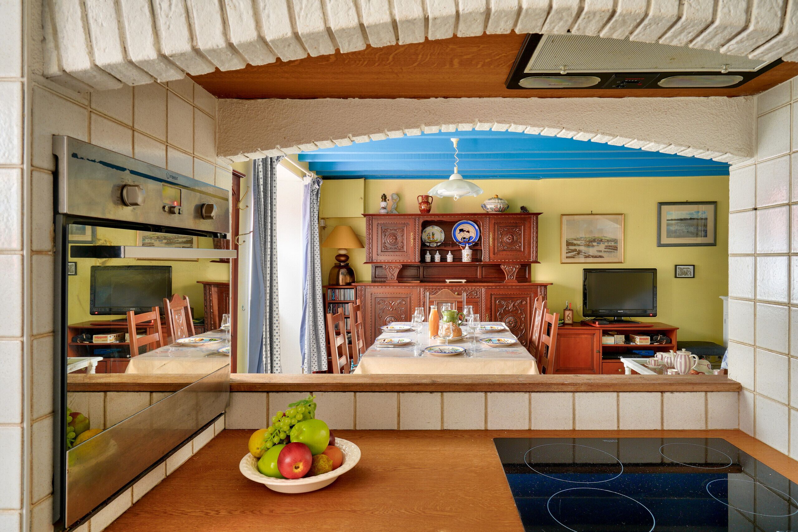 A kitchen with a dining area, a built-in oven, a television, and a fruit bowl on the counter.