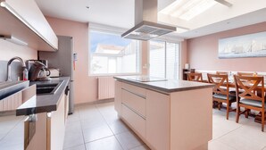 A modern kitchen with a central island, a dining area with wooden chairs, and a large window providing natural light. - House for 8 – Les Sables – 950m from beach (Les Sables-d´Olonne)