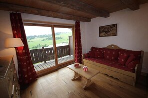 Une pièce avec un plancher en bois, un canapé avec des coussins rouges, une table ornée d’un objet rouge et blanc, et une vue sur un paysage verdoyant par une porte vitrée.