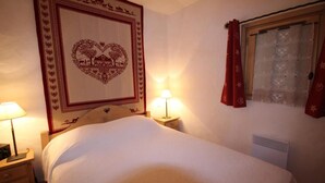 Cama arrumada com um tapete em formato de coração acima, duas luminárias de cabeceira e uma janela com cortinas vermelhas.