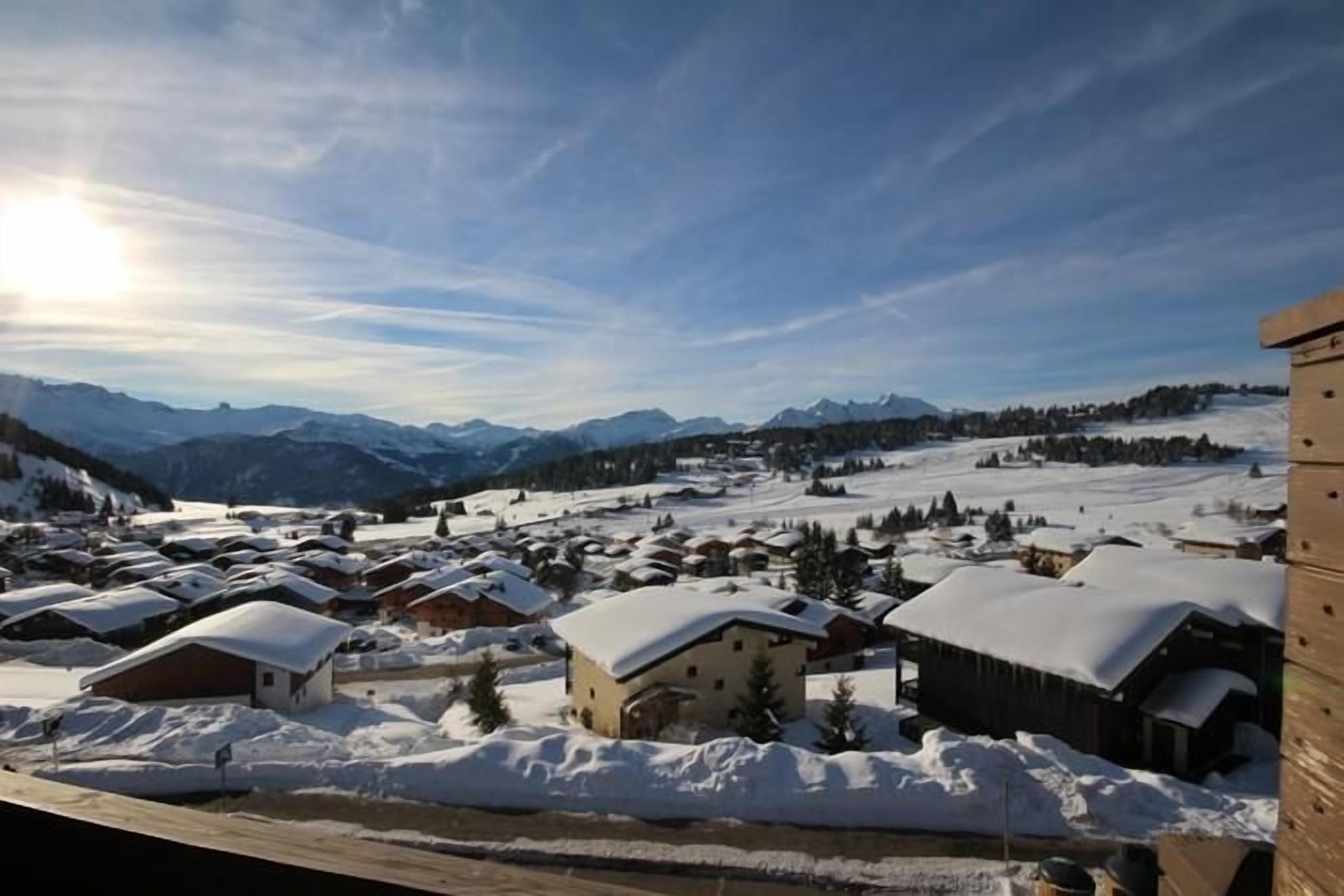 Un village enneigé, avec ses maisons traditionnelles en bois, un ciel dégagé et des montagnes en arrière-plan.