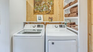 A laundry room with two front-loading washing machines, a wooden wall with a tapestry, and shelves with folded towels. - Tranquil Fairmont Studio w/ Patios - Pets Welcome! (Fairmont)