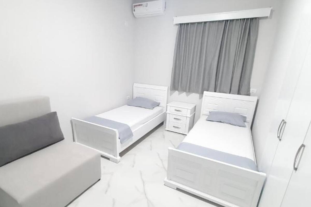 A modern bedroom with two single beds, a grey sofa, a wardrobe, and a window with grey curtains.