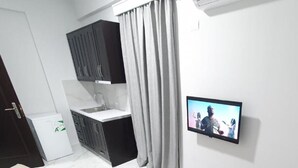 Un saló modern amb un sofà, una taula petita, un televisor fixat a la paret, una cuina amb armaris i una finestra amb cortines.