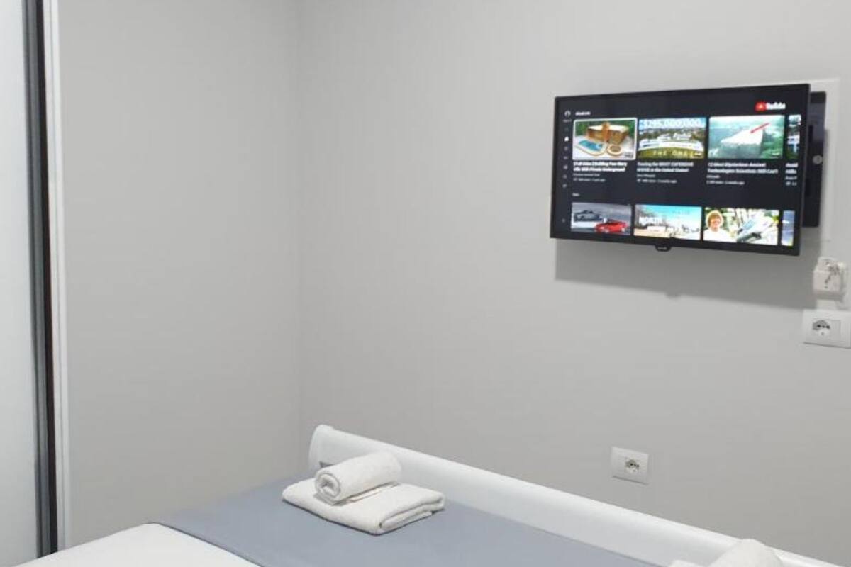 A hotel room with a bed, a small table, and a wall-mounted TV displaying various channels.