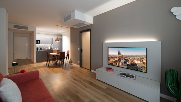 A modern living room with a flat-screen TV mounted on the wall, a red sofa, and a dining area with a table and chairs.