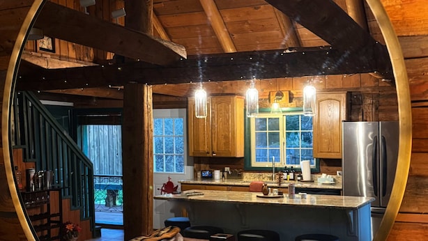 Un espejo circular que refleja el interior acogedor de una cabaña con techo de madera, una zona de cocina y un sofá de cuero.