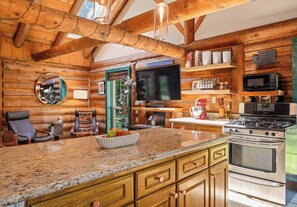 L’intérieur d’une cabane en rondins comprend un espace cuisine doté de plans de travail en granit, d’armoires en bois, d’une cuisinière et d’un téléviseur.