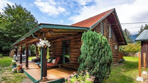 Una cabaña de troncos con un porche, un área de estar cubierta y un gran árbol de hoja perenne.