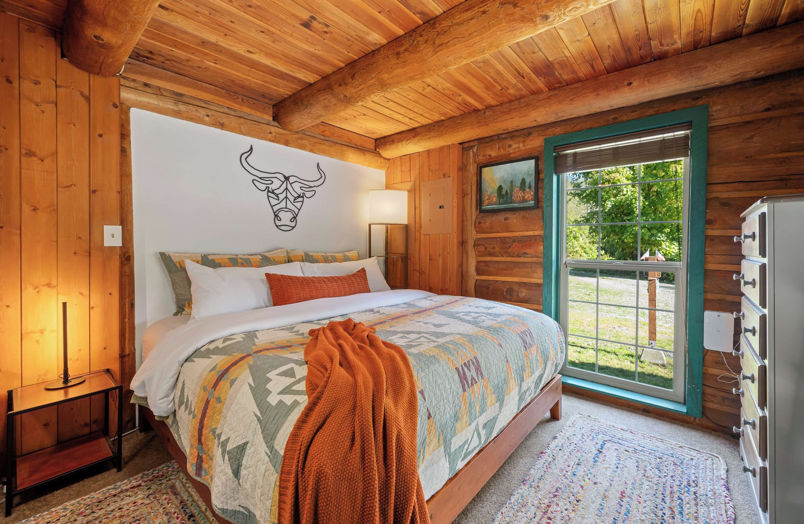 Un dormitorio tipo cabaña de madera con cama, decoración de cabeza de toro en la pared, ventana con vistas y cómoda.