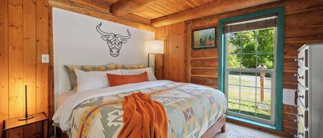 A wooden cabin bedroom with a bed, a wall-mounted bull head decoration, a window with a view, and a dresser.