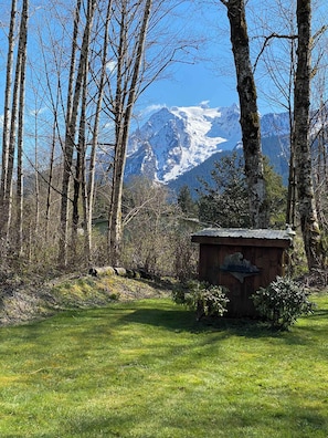 Una cabaña de madera con techo metálico, rodeada de árboles y un área verde, con una cadena montañosa al fondo.