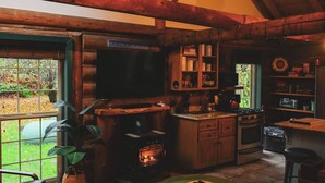 Interior rústico de una cabaña con chimenea, vigas de madera, televisor y zona de cocina.