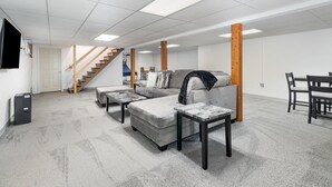A spacious living room with a grey sectional sofa, two ottomans, a coffee table, and a dining table with chairs. A staircase with wooden railings leads to an upper level. - Hershey's Hideaway: A Tranquil Retreat on the Hill (Palmyra)