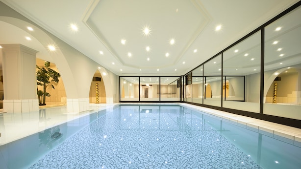 A large indoor swimming pool with a tiled floor, surrounded by glass walls and white columns. - Das Kronberg - Adults Only (Bodenmais)