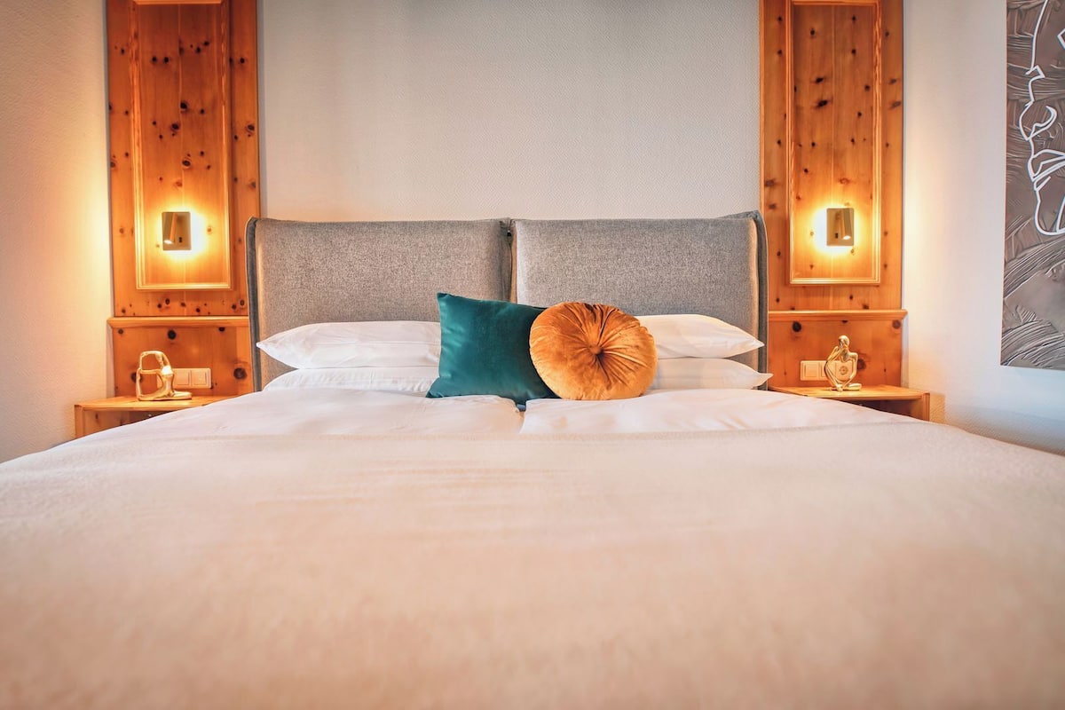 Ein ordentlich bezogenes Bett mit weißen Leinentüchern, zwei Kissen und einem dekorativen Rundkissen. An den Holzkopfteilen befinden sich Wandleuchten.
