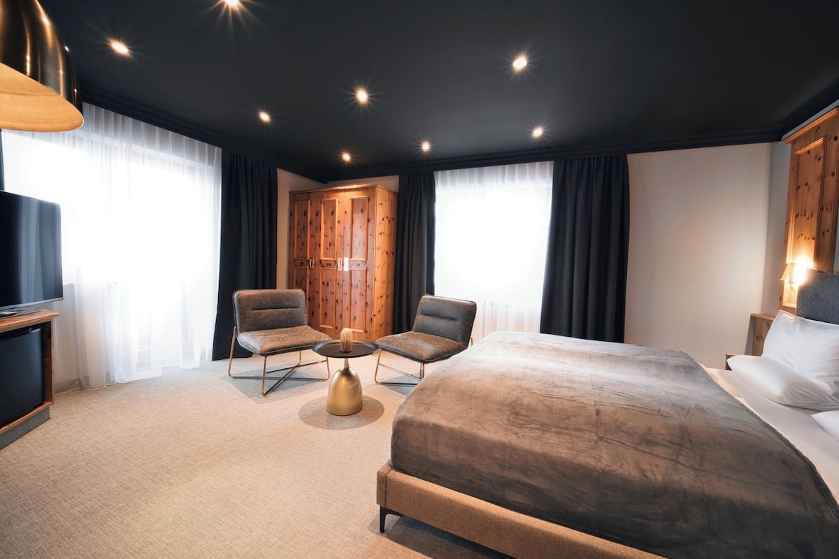 Ein modernes Hotelzimmer mit einem großen Bett, zwei Sesseln, einem kleinen runden Tisch und einem Fernseher.