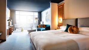 A modern hotel room with a large bed, a seating area with a blue chair, a small table, and a view of the outdoors through a large window. - Das Kronberg - Adults Only (Bodenmais)