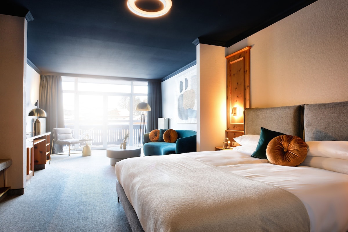 Ein modernes Hotelzimmer mit einem großen Bett, einer Sitzecke mit blauem Sessel, einem kleinen Tisch und Blick ins Freie durch ein großes Fenster.
