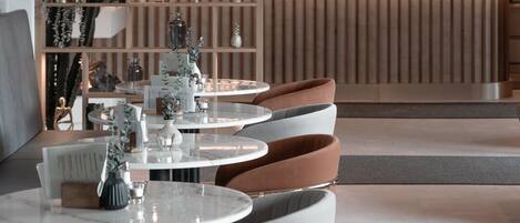 A modern dining area with round tables and contemporary chairs, a decorative hanging light fixture, and a background featuring a wooden panel wall with shelves displaying various items.