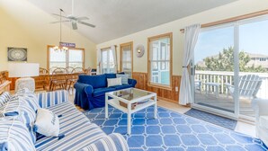 A spacious living room with a blue patterned carpet, a blue sofa, a coffee table, and a ceiling fan. There is a dining area with wooden chairs and a table, and a large window with curtains. - Pet-friendly oceanside Corolla home w/ private pool & hot tub! (Corolla)