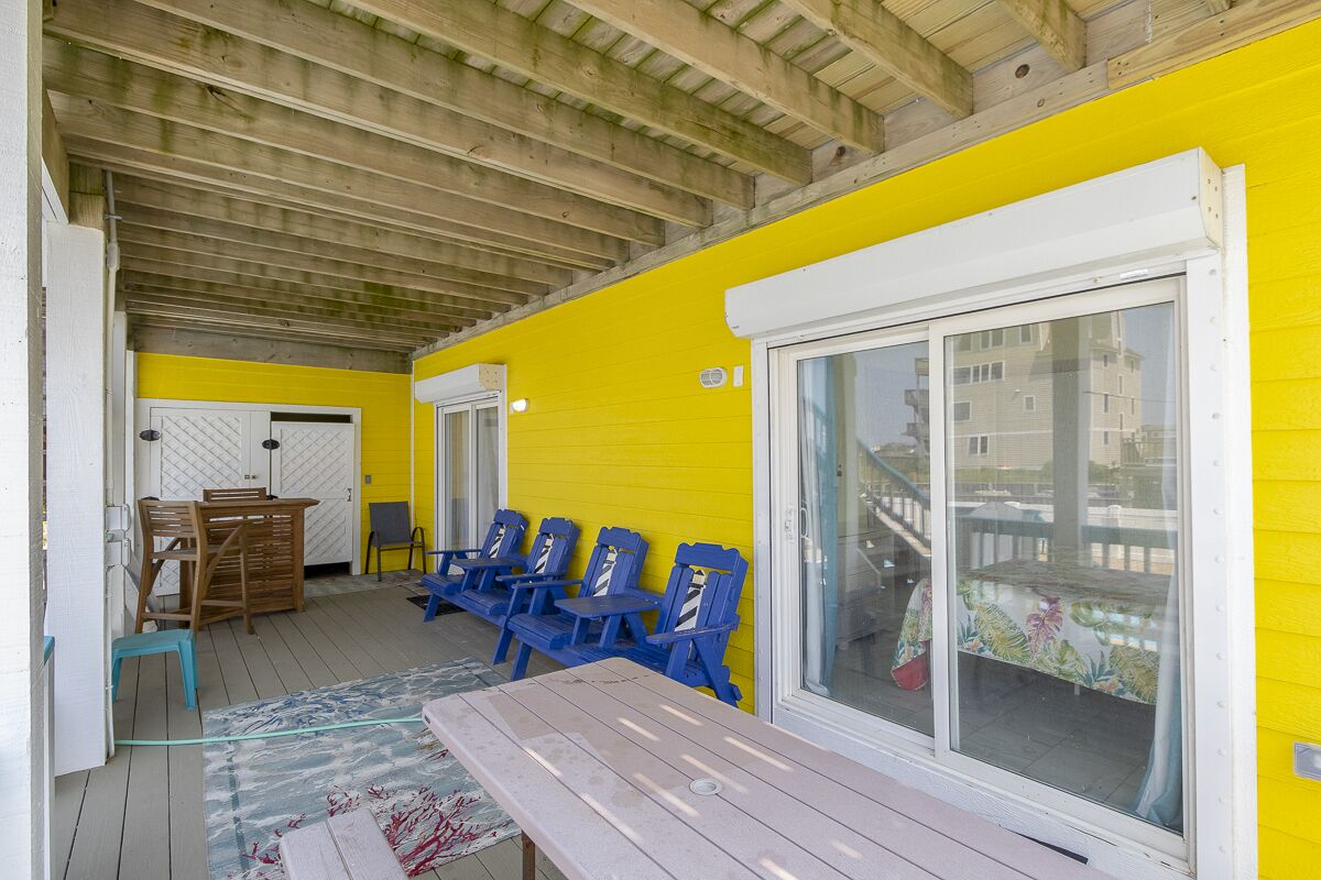 A covered outdoor area with a yellow wall, a large sliding glass door, and blue chairs.